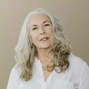  Imagem de uma mulher branca com cabelo longo e branco, usando uma camisa branca sobre um fundo bege escuro